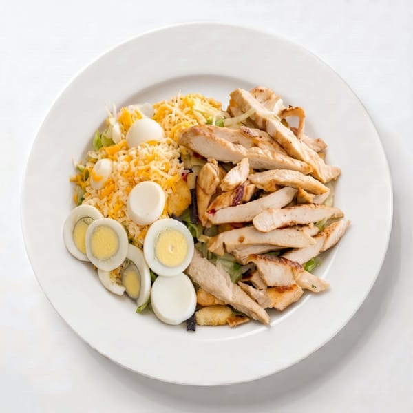 Grilled chicken salad with shredded cheese, hard-boiled eggs, and croutons on a white plate.