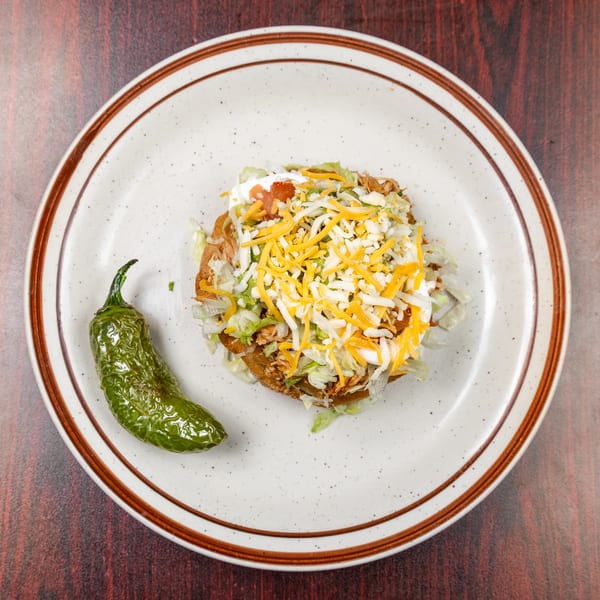 Tostada with shredded meat, lettuce, cheese, and sour cream, served with a green pepper on a plate.