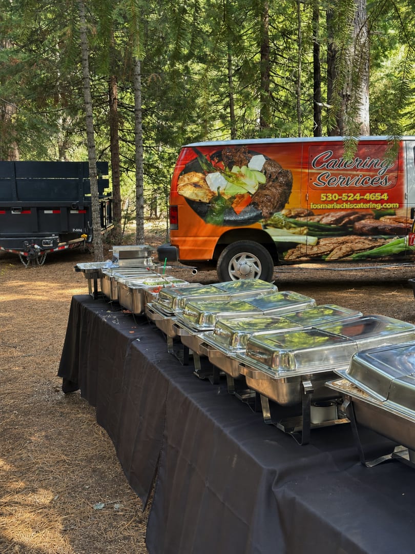 Catering buffet with chafing dishes on a black-draped table, with a catering van in a forest.
