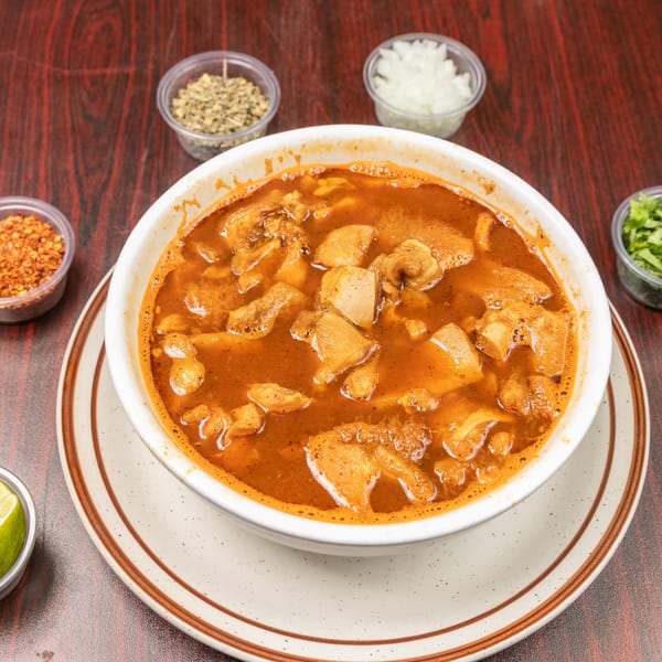 Bowl of menudo with tripe, served with lime, onions, cilantro, chili flakes, and oregano.