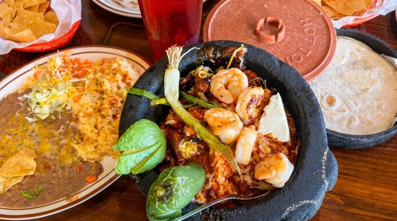 Shrimp molcajete with rice, refried beans, tortillas, and chips on a wooden table.