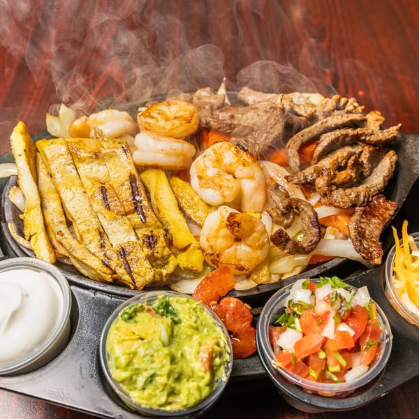 Steaming fajita platter with steak, chicken, shrimp, vegetables, and sides of guacamole, pico, and cheese.